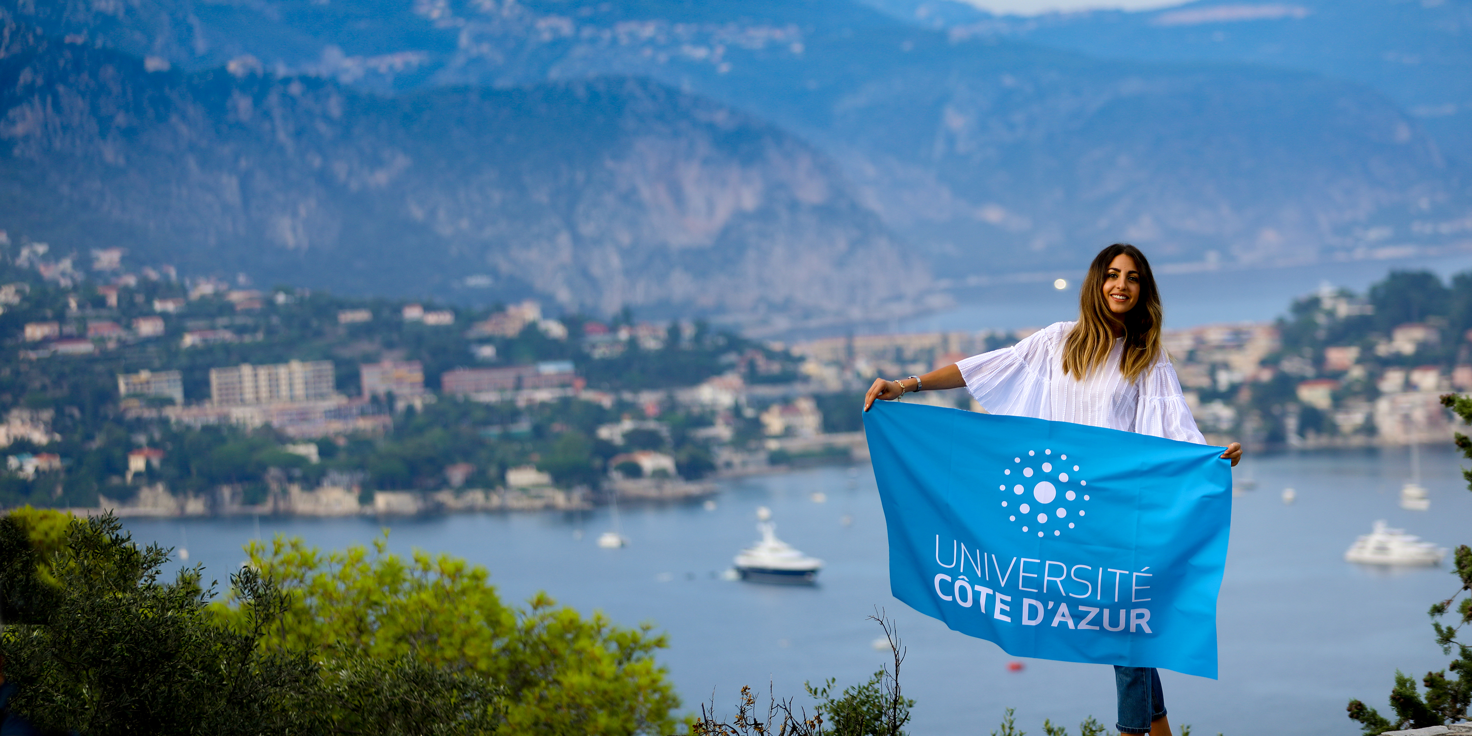 Jeaune femme tenant le drapeau UCA devant un paysage de mer et montagne